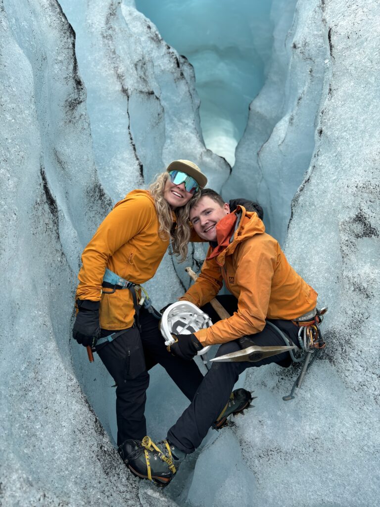 Two smiling owners of Odyssey posing at the entrance of a light blue ice cave on Solheimajokull glacier.